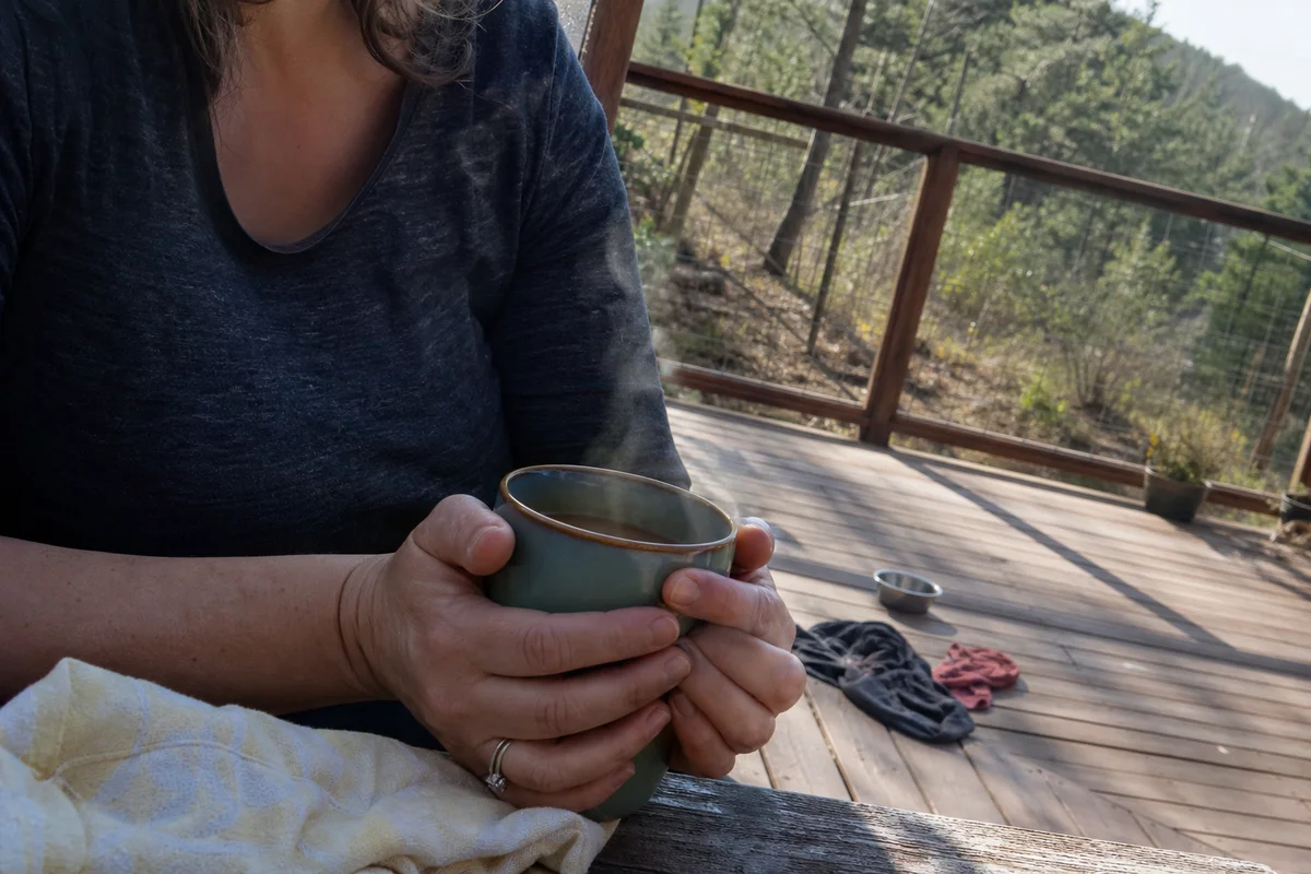 Hands wrapped around a warm mug on a quiet deck, forest beyond, sense of pause