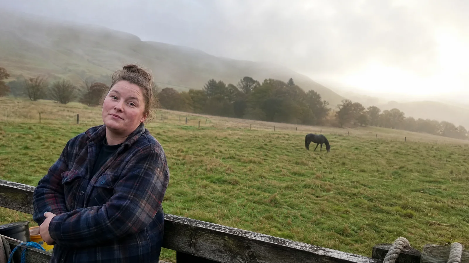 Woman resting against a paddock fence at dawn, tired but softening, horse grazing in the background, gentle fog